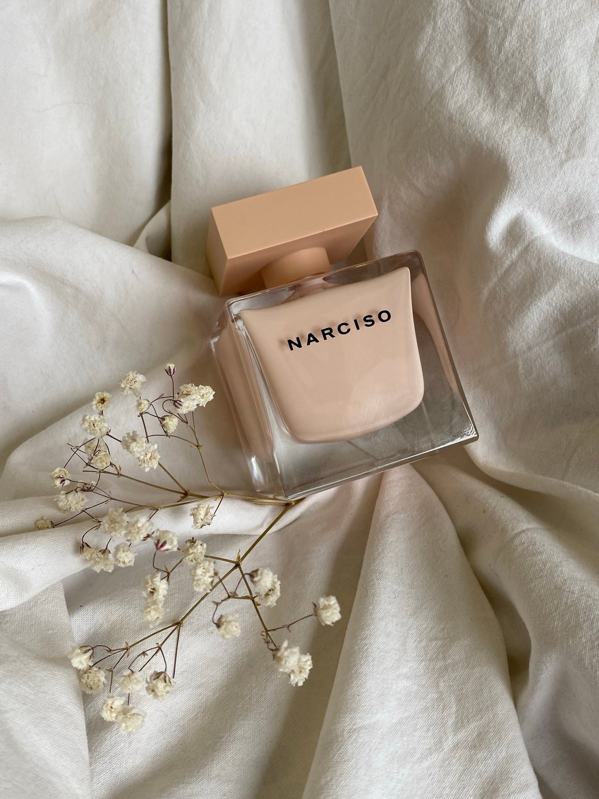 Square perfume bottle resting on white linen with delicate dried flowers.
